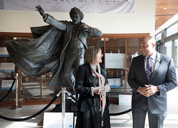 Christine Kinealy and Don Sawyer gesture in front of a statue of Frederick Douglass