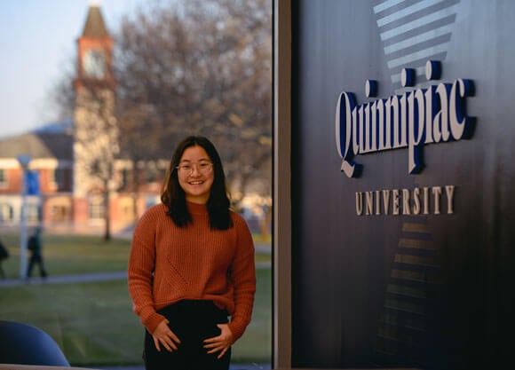 Ashley Hong smiles and poses in front of Quad and next to Quinnipiac signage on wall.