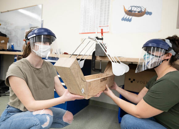 School of Engineering students Madeline LePage (left) and Gabriella Ambery work on their senior projects while wearing face shields and masks and holding a block of wood.