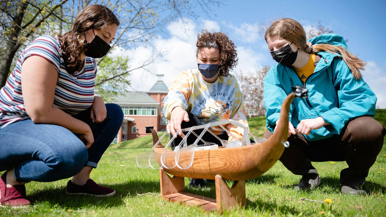 Three students sit in the grass on campus with a wooden row boat in the center.
