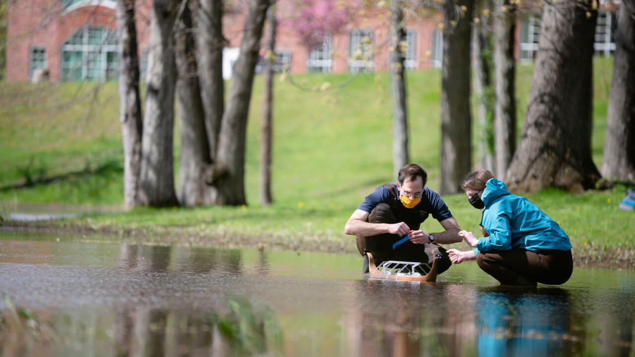 A student and professor kneeling down near a pond testing the wooden row boat.