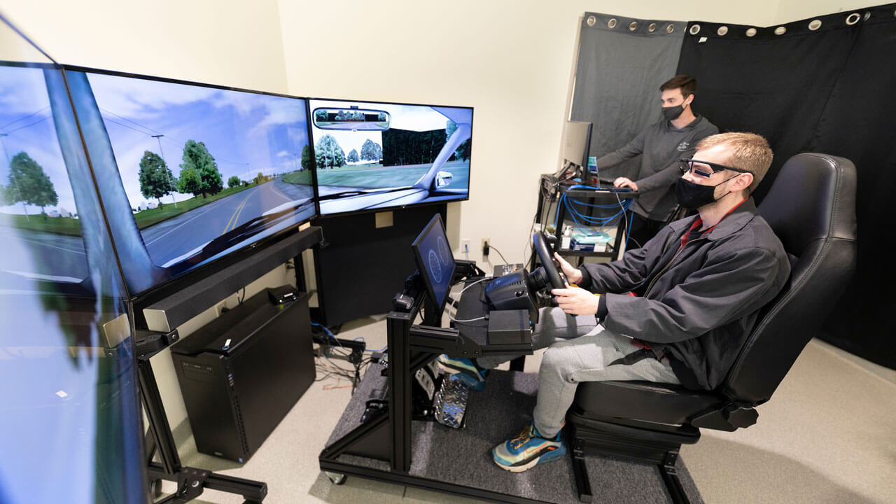 A student sitting in a chair holding a steering wheel using a driving simulator looking at three large screens.
