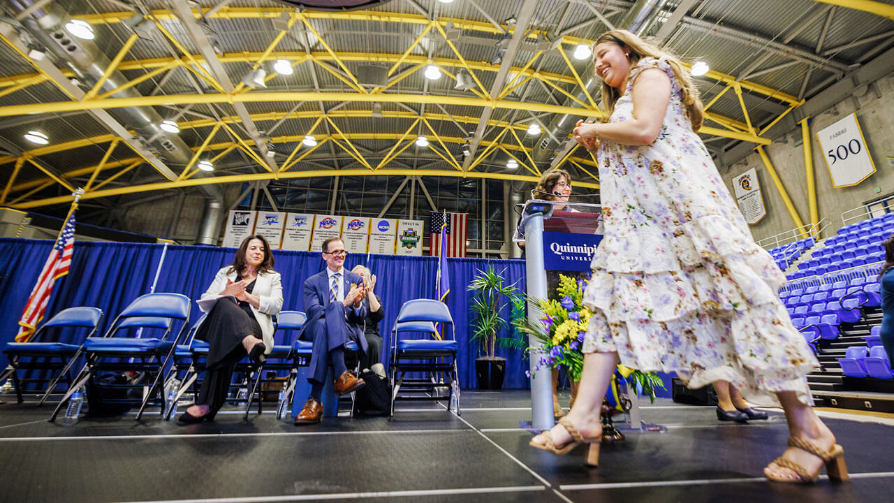 blonde female student in a long floral dress walks across the stage in heels