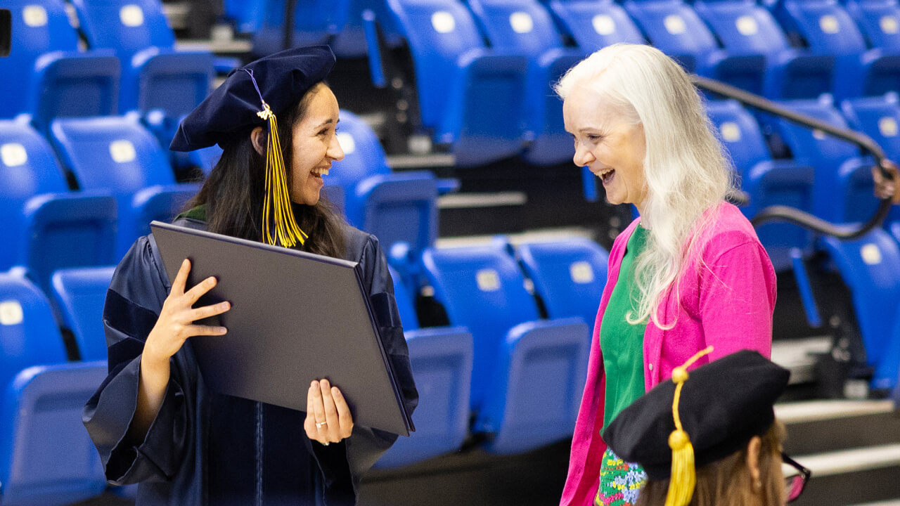 A graduate smiles as she shows her guest her diploma