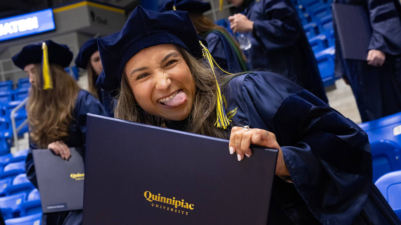 A graduate cheerfully sticks out her tongue and holds her diploma up as she poses for the camera