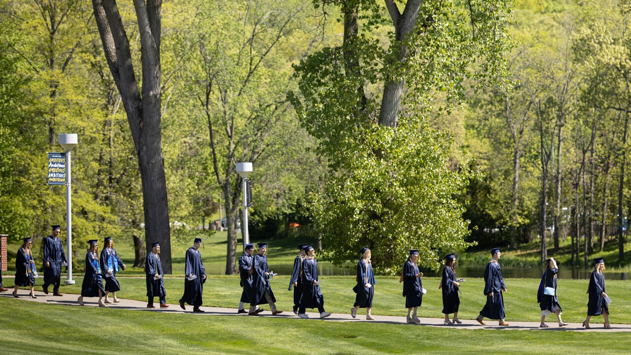 A dozen graduates in the distance walk on a path across the quad under a sign that reads Ambition