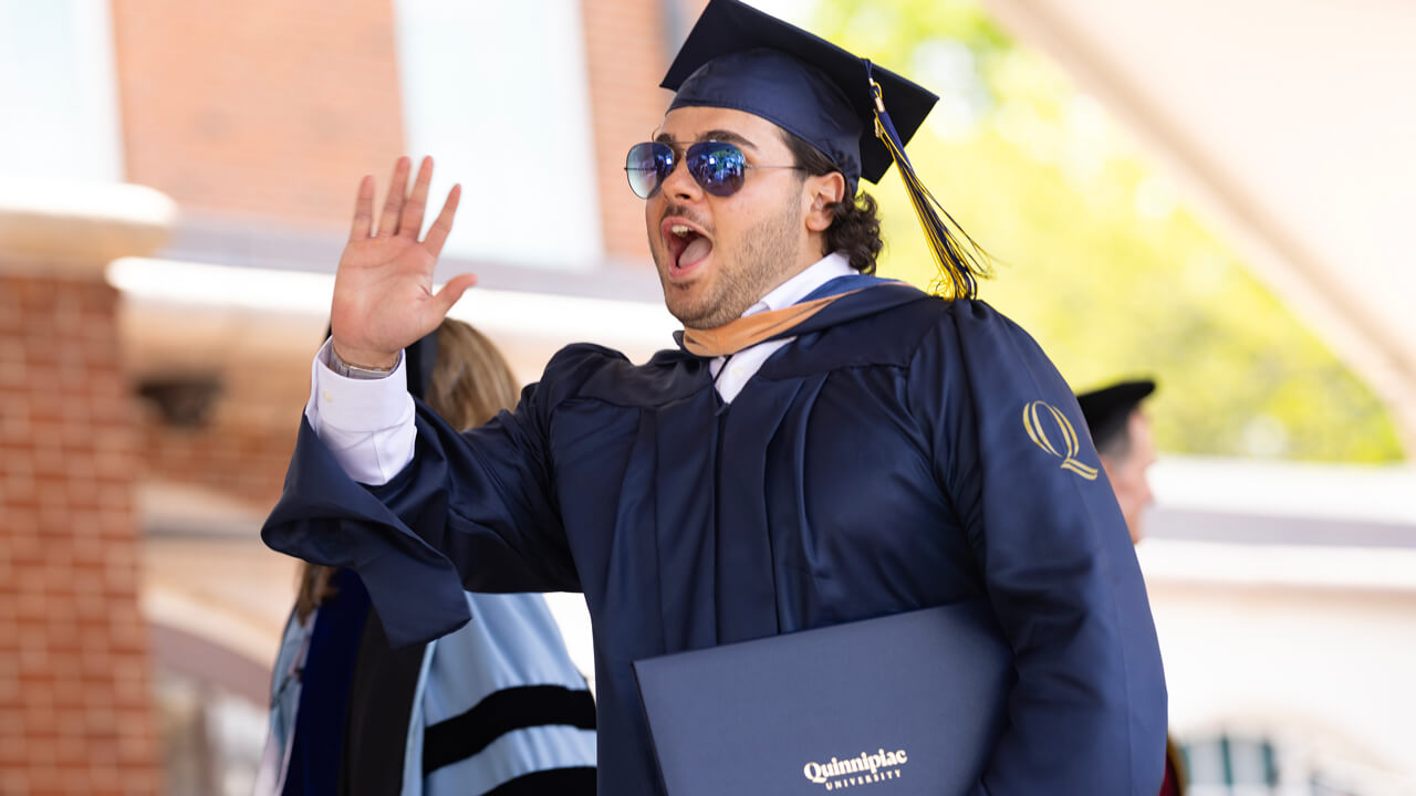 A graduate wearing blue sunglasses holds his diploma under his arm and waves