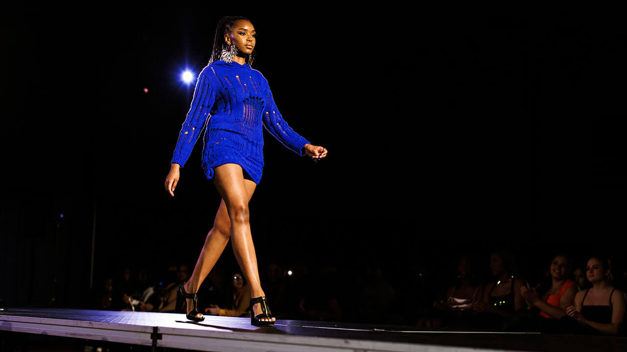 woman with long braided ponytail walks down the runway in black heels and a knit blue dress