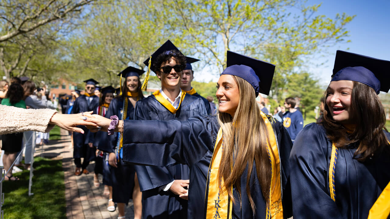 A graduate shakes hands with an audience member as she walks down the path on the quad in line with other graduates