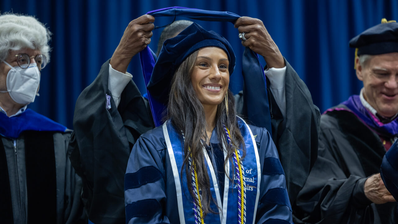 A graduate smiles as a faculty member places a doctoral hood over her head