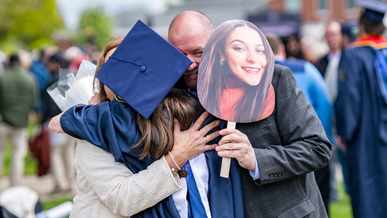 A graduate's family member cries and holds a sign with the graduate's face as he hugs her
