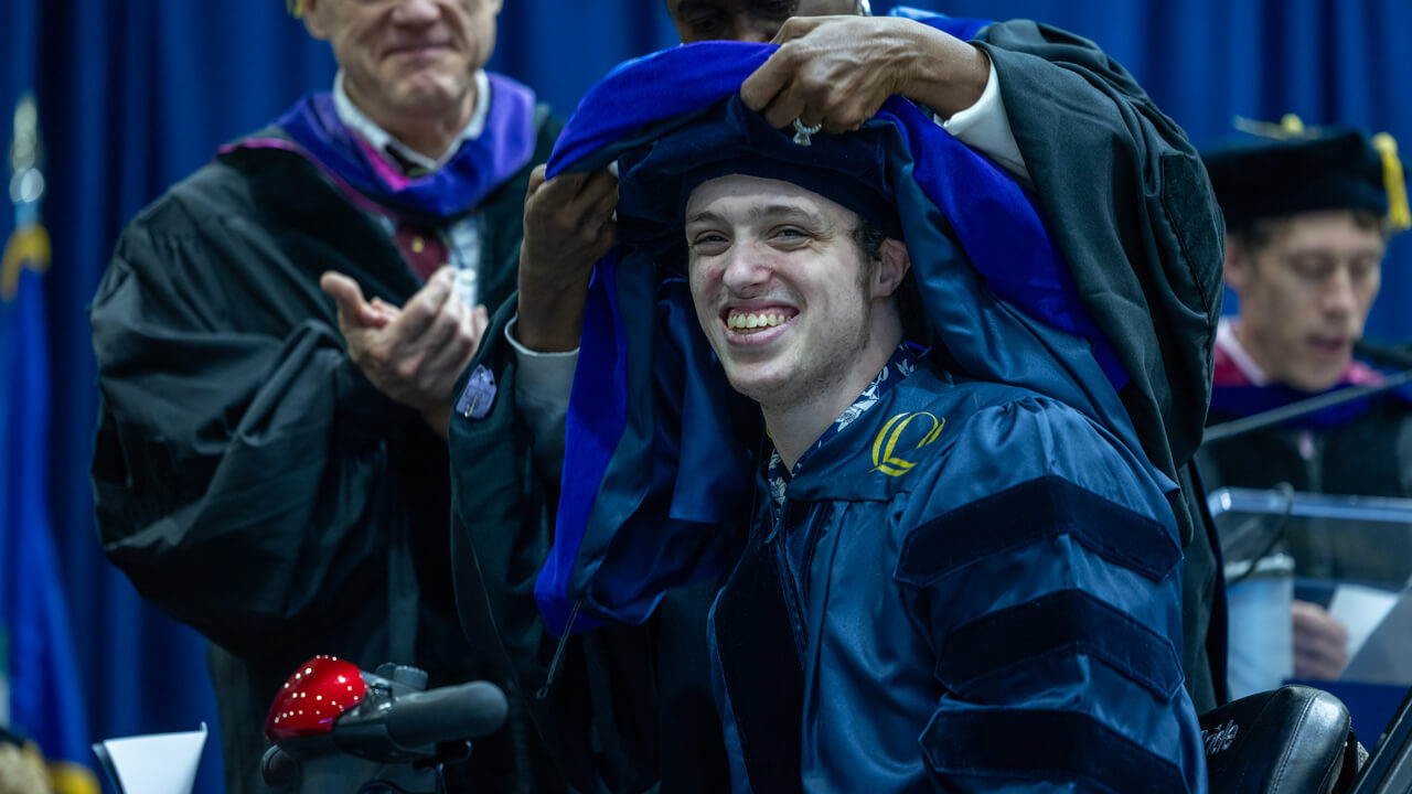 A graduate smiles broadly while a professor lifts a doctoral hood over his head