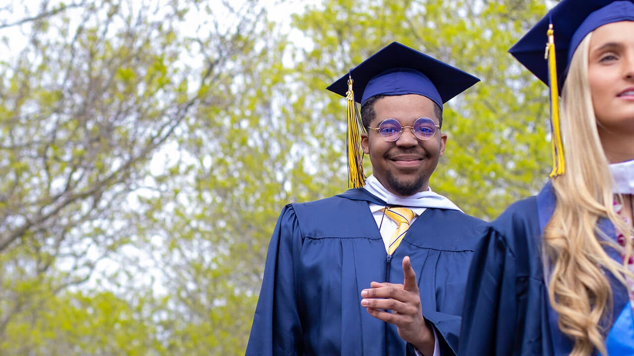 A graduate in shiny blue sunglasses points to the camera and smiles