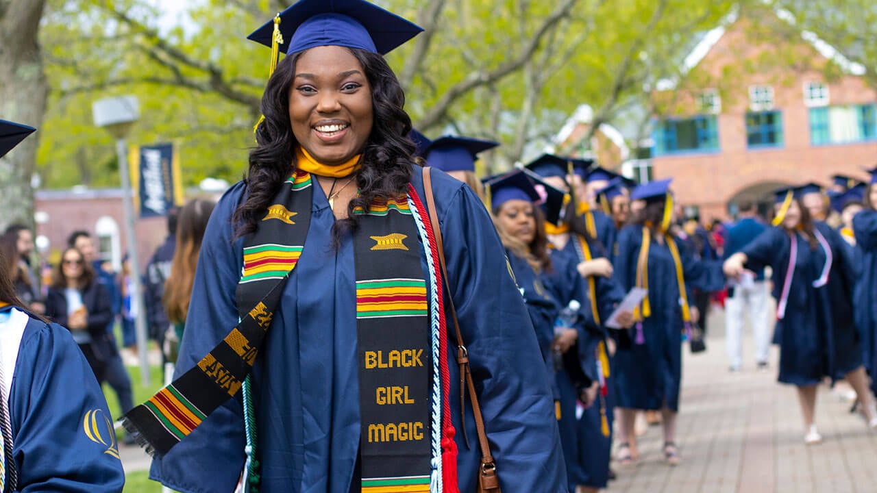 A graduate wearing a stole that reads "Black Girl Magic" walks down a brick path
