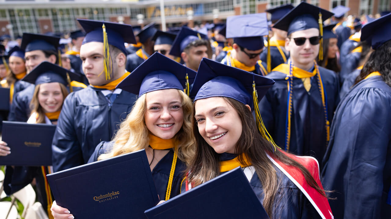 Two grads pose for a photo with their diplomas among a sea of graduates