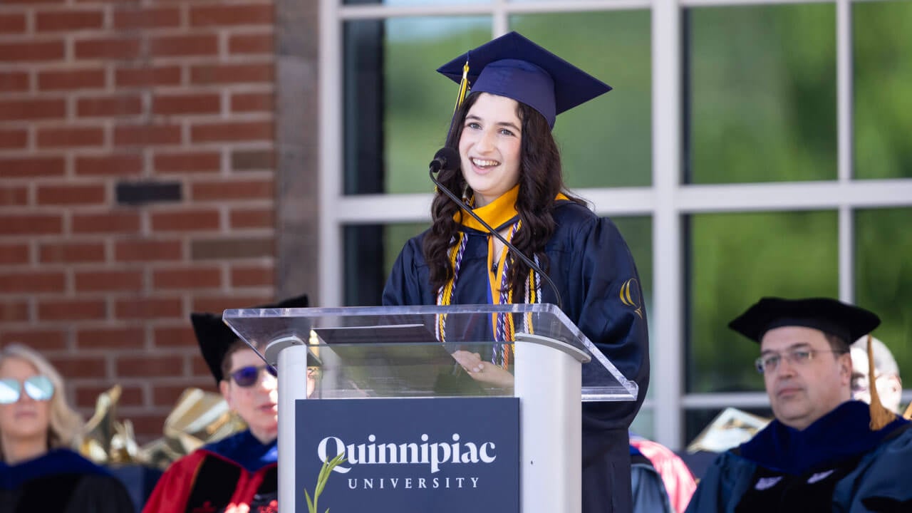 Student stands at the commencement podium in cap and gown and speaks into the microphone