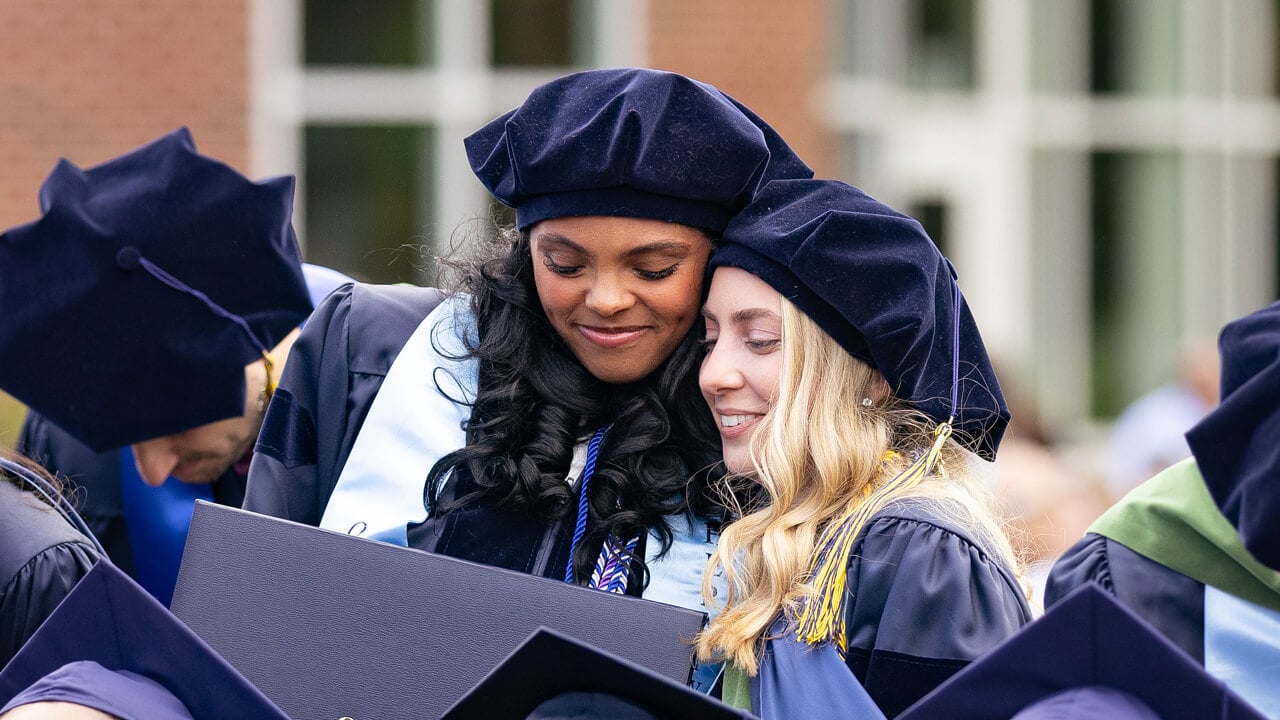Two graduates embrace warmly as they hold their diplomas