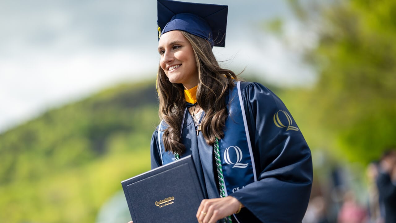 A graduate wearing a navy stole holds her diploma with the Sleeping Giant in the background