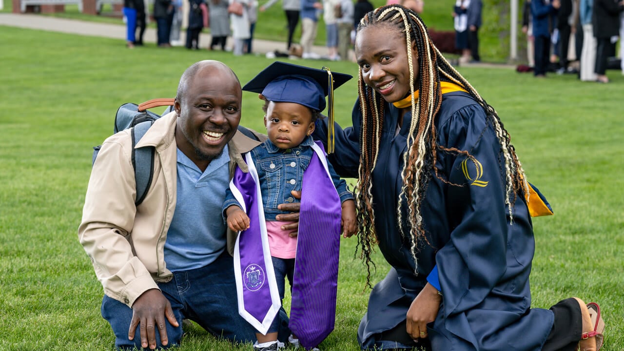 A graduate kneels down for a photo with her guest and a toddler wearing a graduation cap and stole