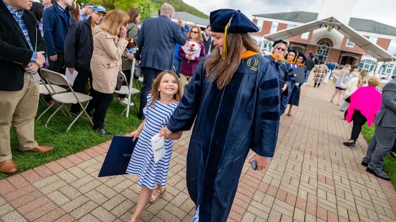 A graduate holds hands with a young girl who lovingly gazes up at her as they walk across the quad