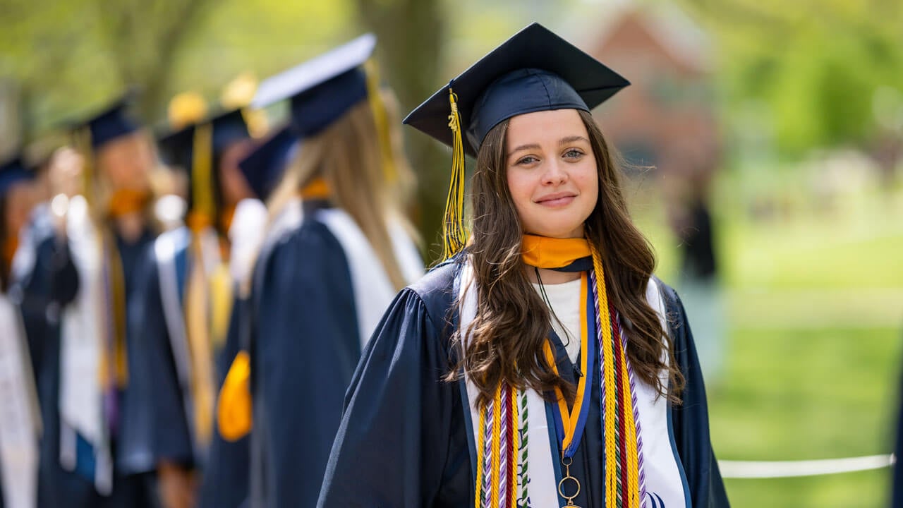 A 2024 Nursing graduate dons a vast amount of academic regalia while standing in line amongst their peers.