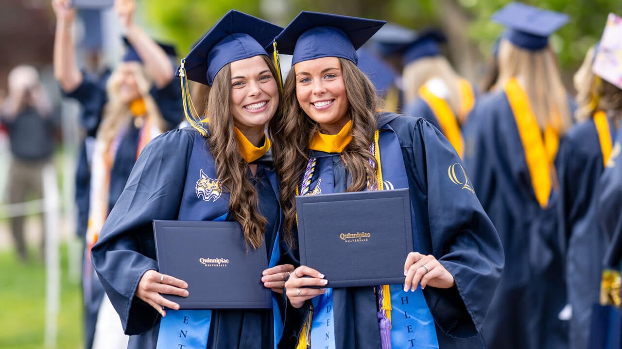 two graduates pose together with their diplomas
