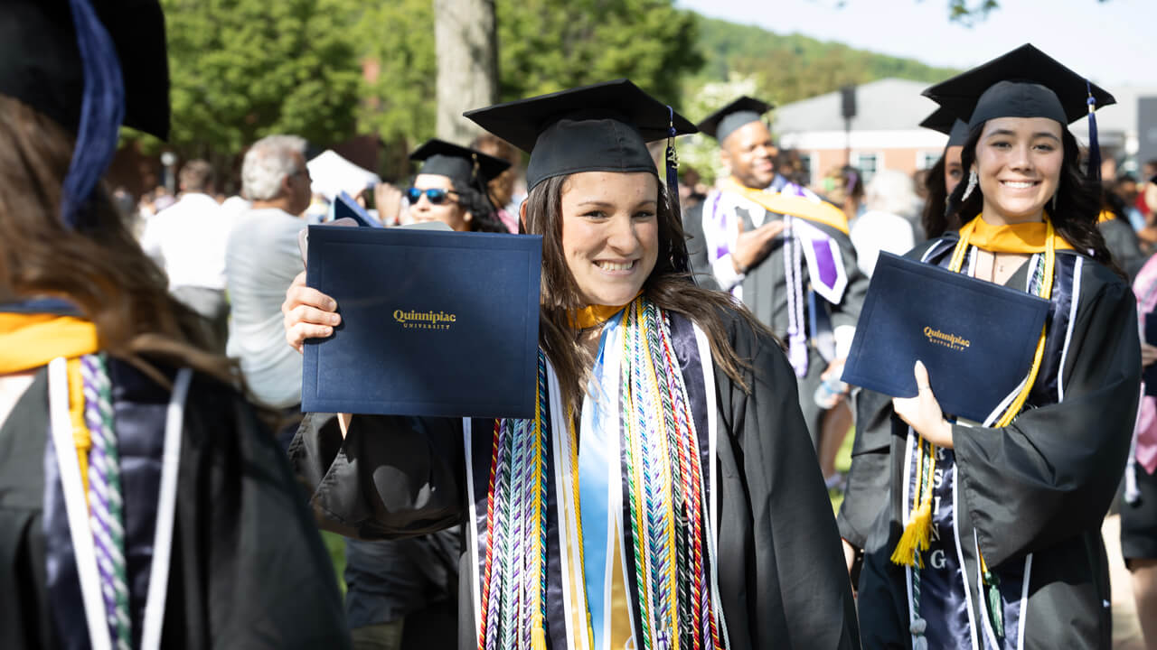 A graduate wearing dozens of honor cords holds her diploma and smiles