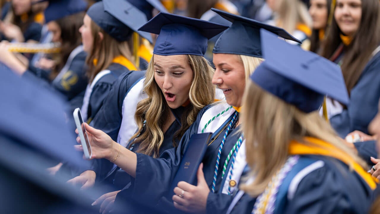 Two graduates look shocked at something shown on one of their phones as they sit with their degrees.