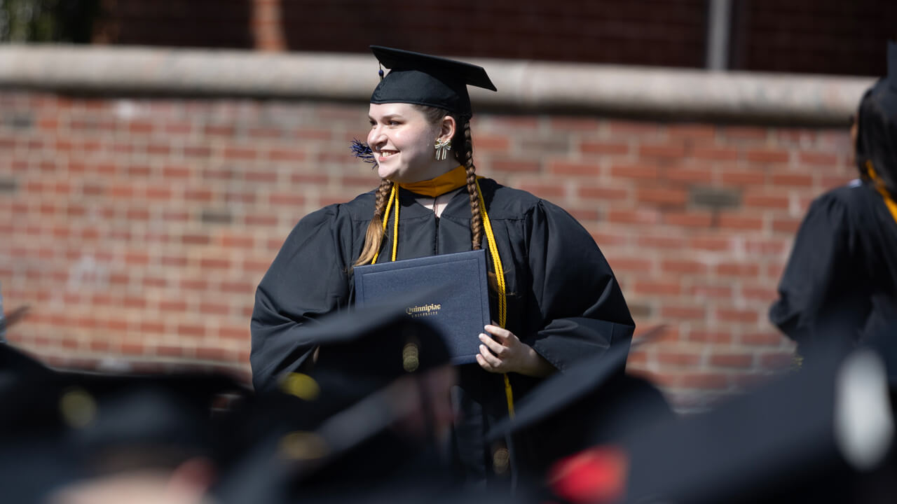 A graduate stands and smiles with her diploma behind dozens of other graduates