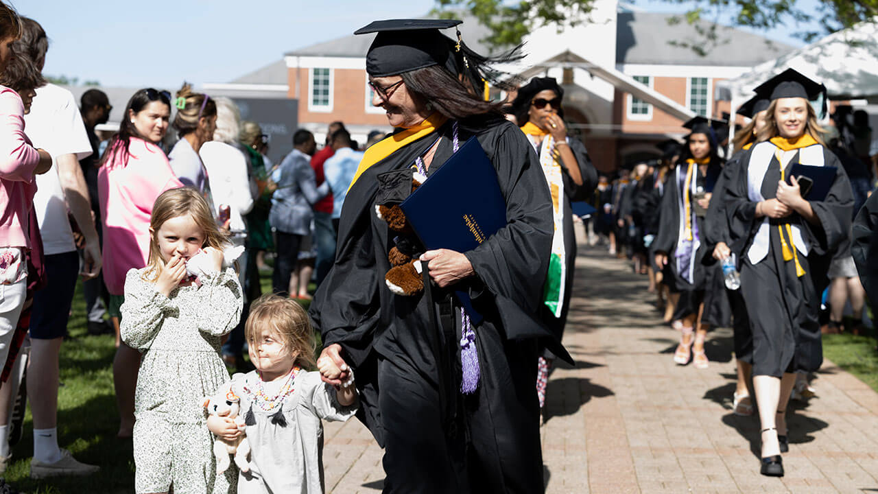 graduate walks down the aisle holding the hand of a toddler while another small child also follows her