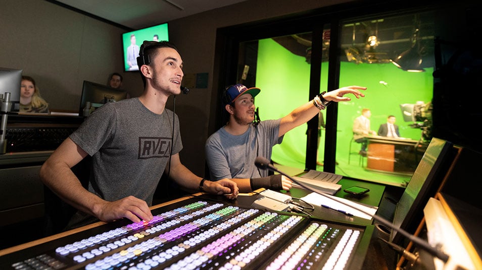 Two students wearing headsets sit behind a large control board in the McMahon Center communications room
