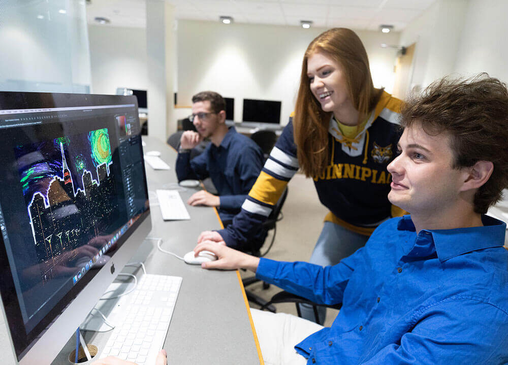 A communications student works on a graphic design on a computer monitor while a classmate smiles over his shoulder