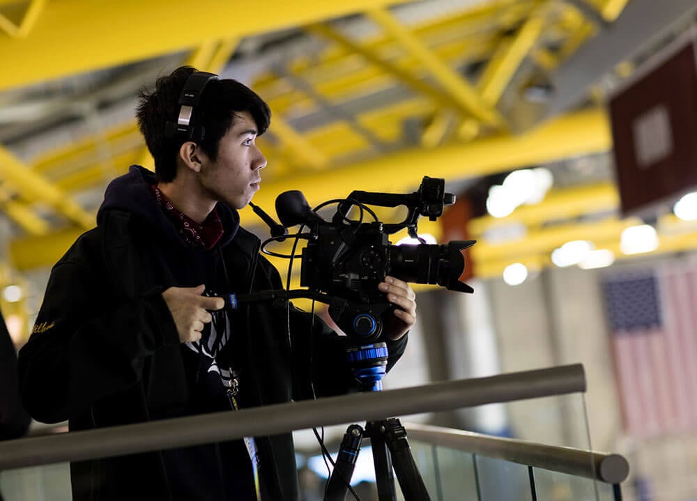 A student wearing headphones and operating a large camera on a tripod films a pregame show at the hockey arena