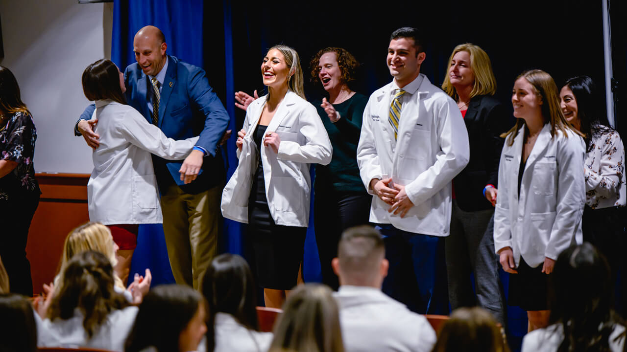 Graduating students are assisted putting on their white coats by faculty members.