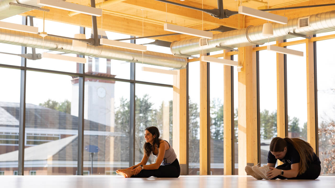 Two students stretch in a studio surrounded by floor-to-ceiling windows