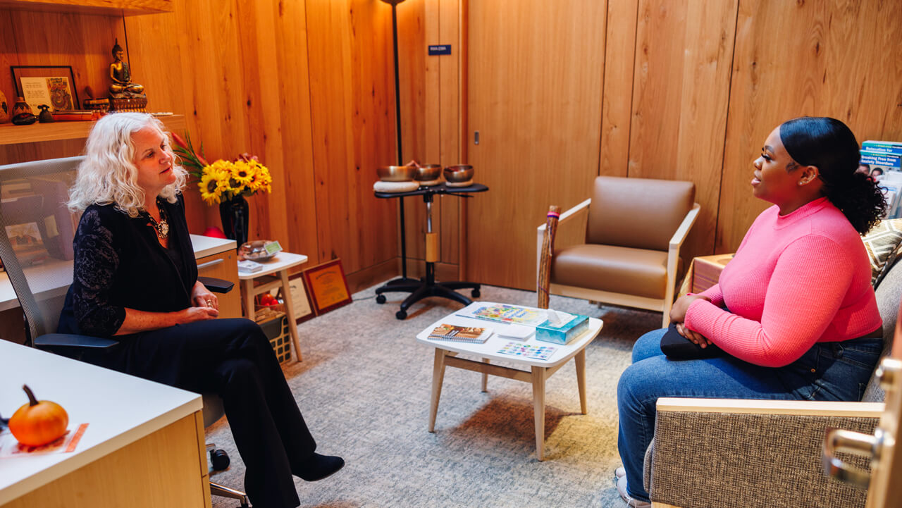 A student and therapist speak together in an office with cozy chairs, natural wood walls and flowers