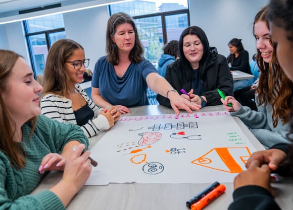 A faculty member and a group of female students stand and sit around a table decorated with health and wellness–related doodles.