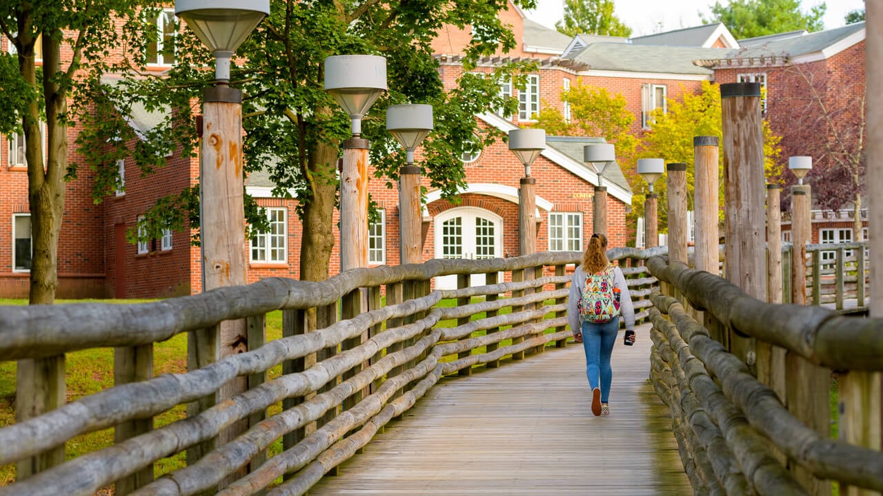 A student walks with a backpack across the Commons bridge toward residence halls