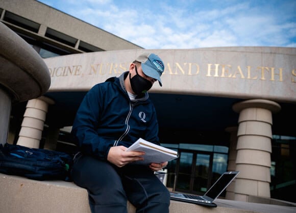 A student wearing a mask sits outside using his laptop