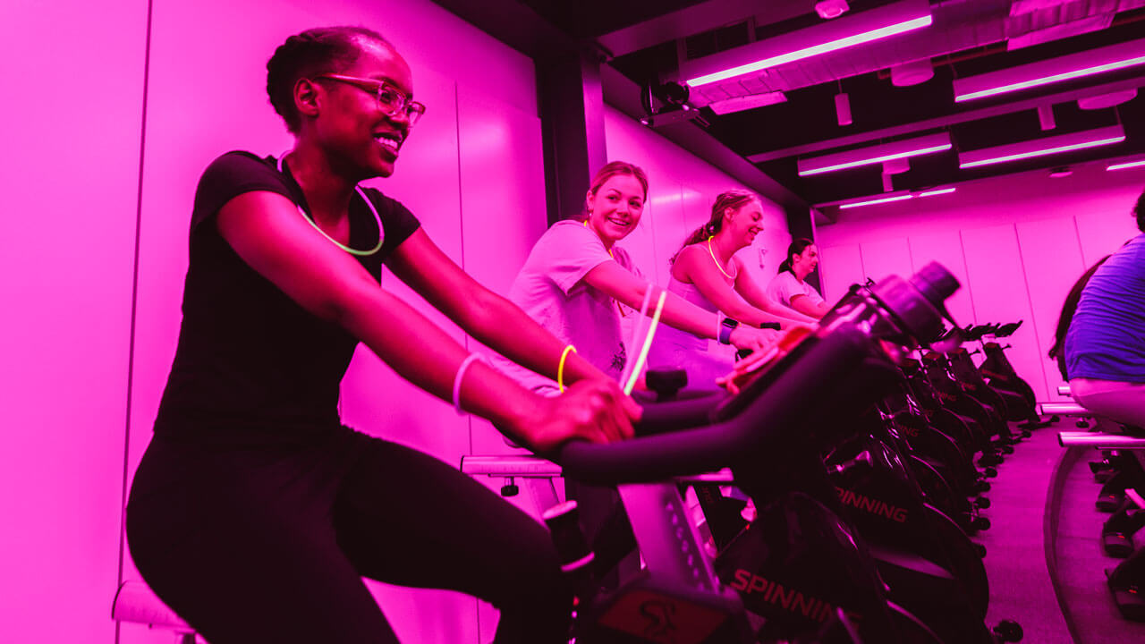 Students ride bikes in a spin class with pink lighting.