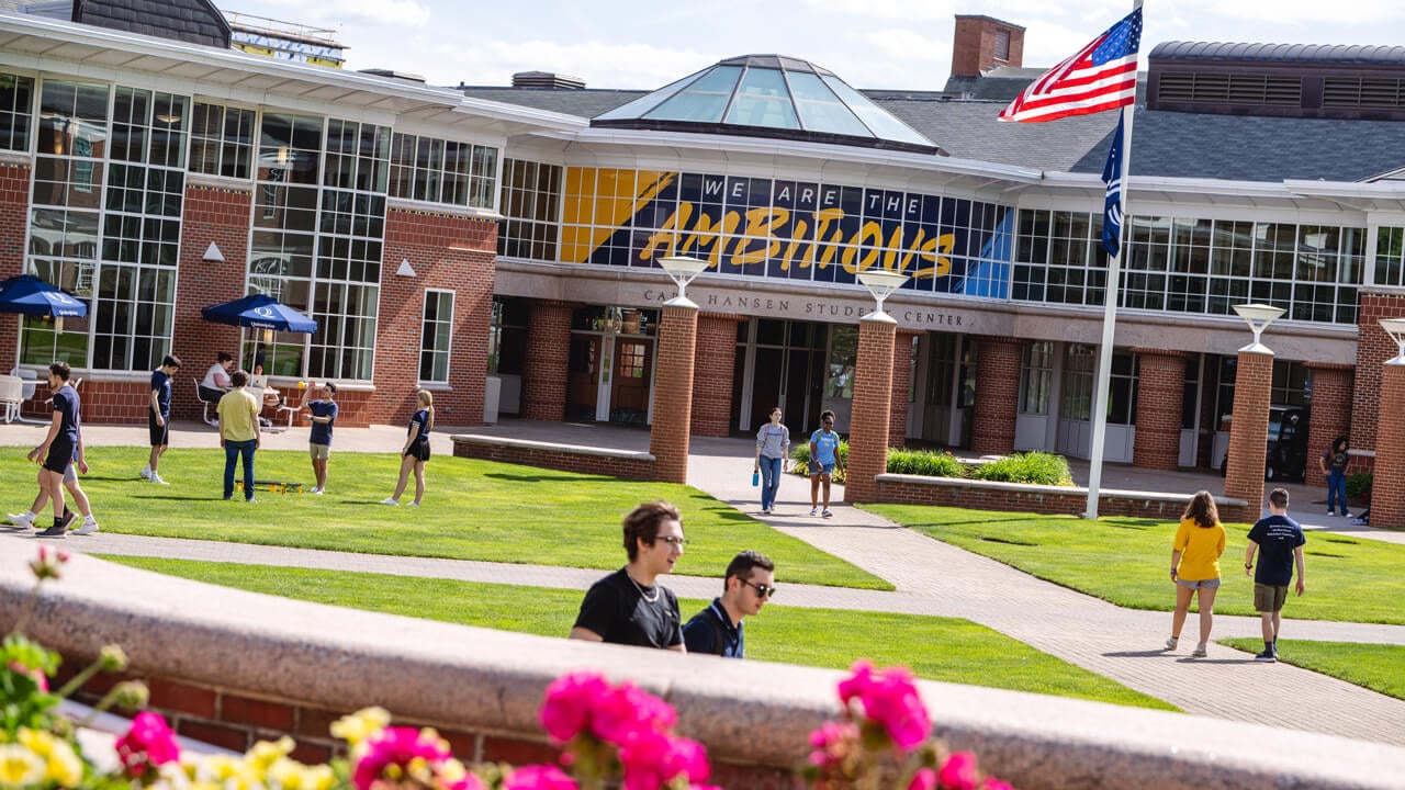Students walk and talk on the lawn in front of the student center with We are the Ambitious in the windows
