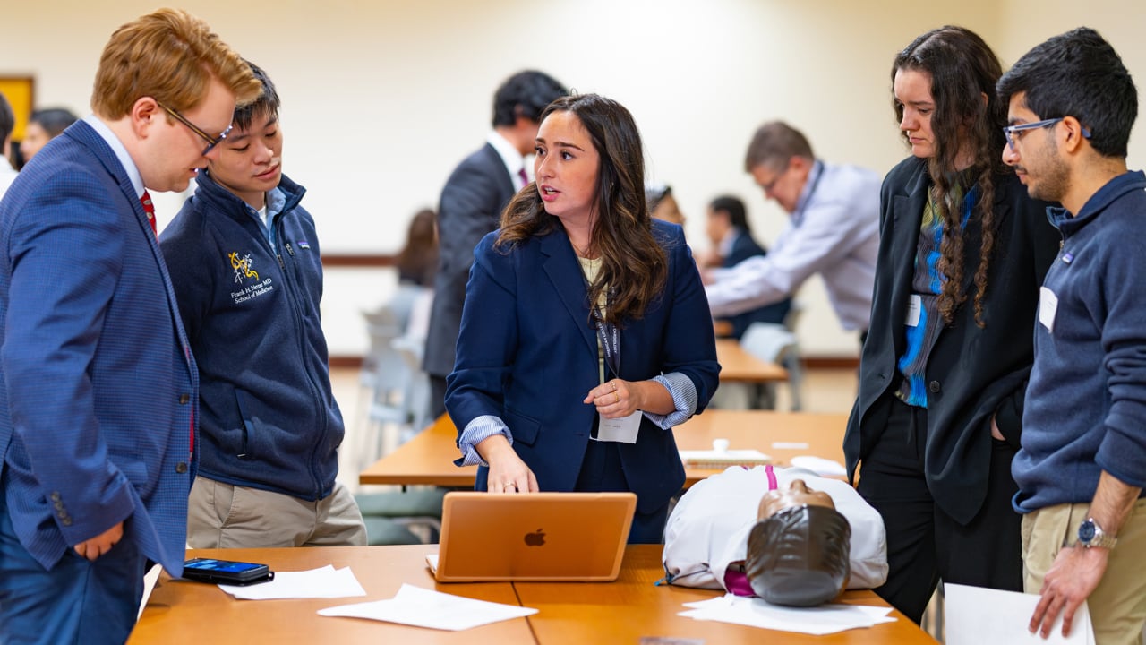 Four students gather around another speaking in front of a laptop.