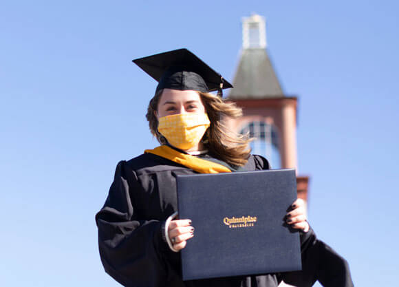 A graduate holds her diploma cover and wears her cap and gown in front of the library