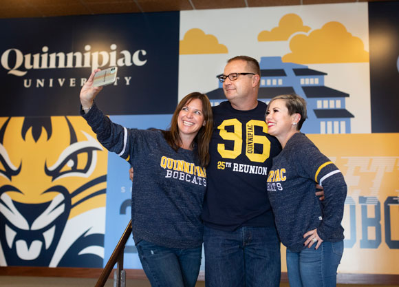 Smiling alumni pose for a selfie in front of a wall with Quinnipiac colors and images