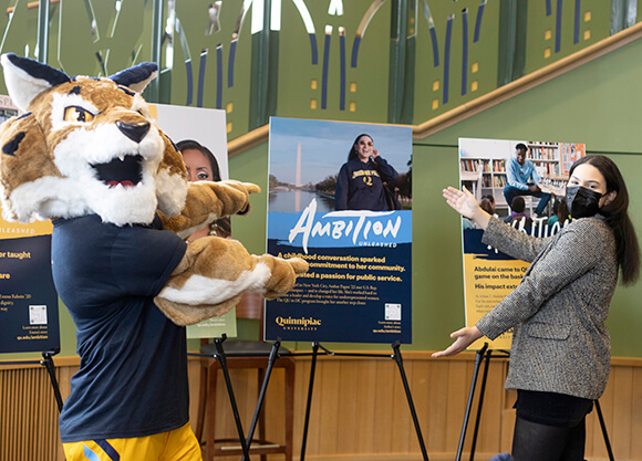 Ambar Pagan and Boomer the mascot celebrate in front of an Ambition Unleashed poster