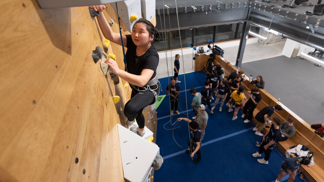 A student hangs onto grips as she climbs the two-story rock wall