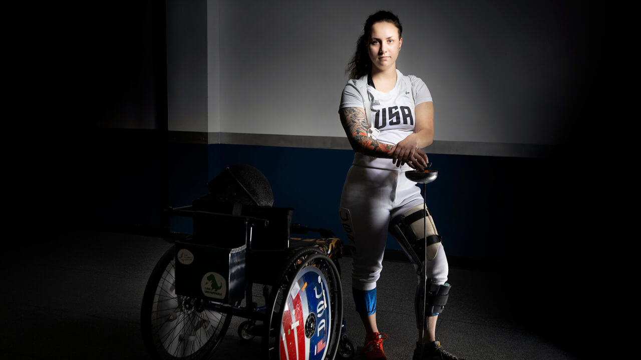 Tori Isaacson wears her USA fencing clothes and stands by her wheelchair holding fencing equipment