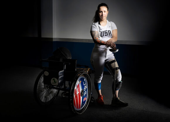 Tori Isaacson wears her USA fencing clothes and stands by her wheelchair holding fencing equipment