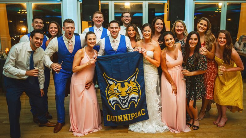 A dozen wedding guests cheer and hold up a large Quinnipiac Bobcats flag