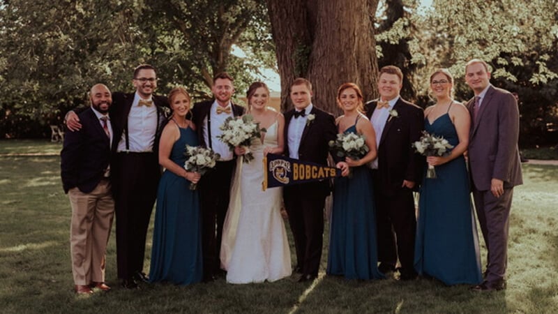 Smiling wedding guests pose under a tree and hold up a Bobcats pennant